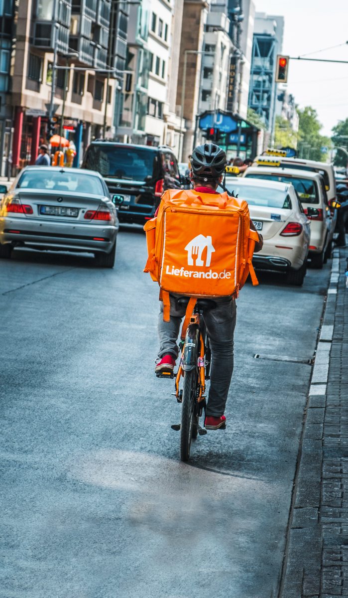 Eine Person auf einem Fahrrad von hinten. Die Person trägt einen orangenen Rucksack eines Lieferdienstes.