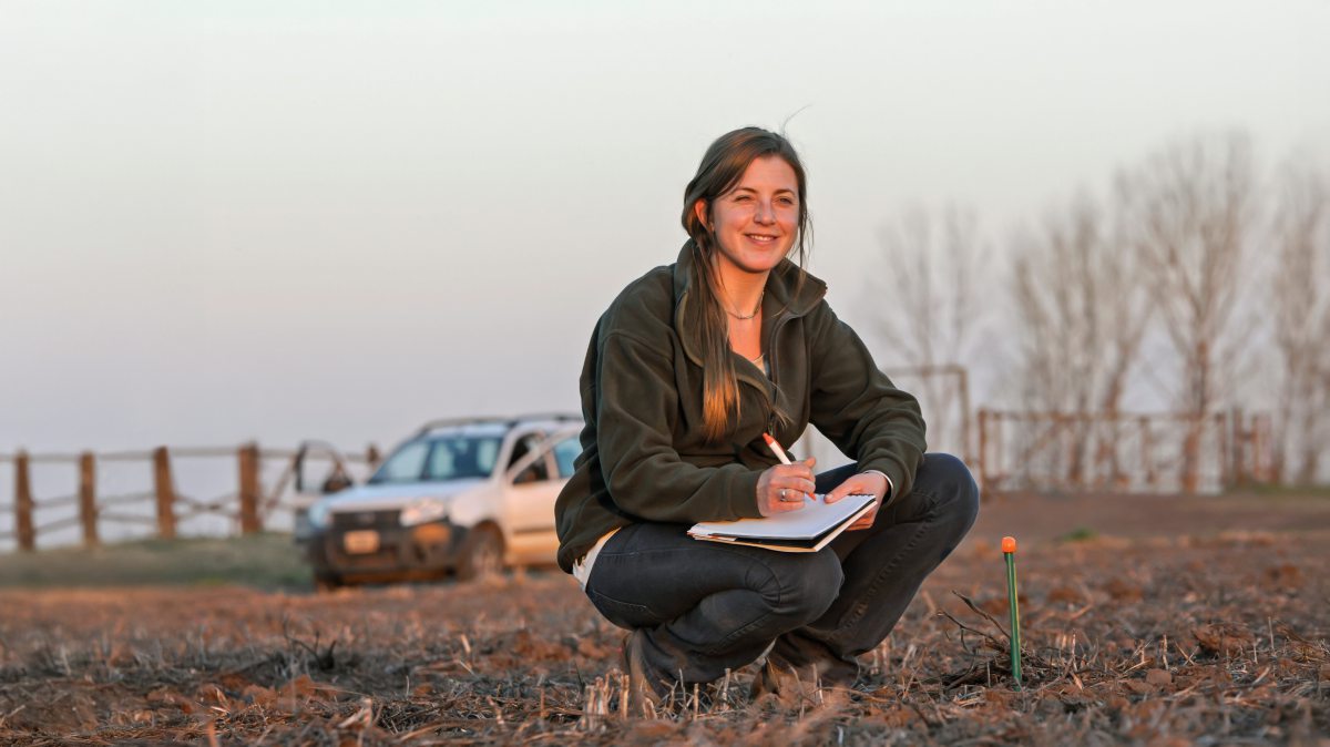 Frau mit Stift und Block hockt auf einem Feld und notiert Daten.
