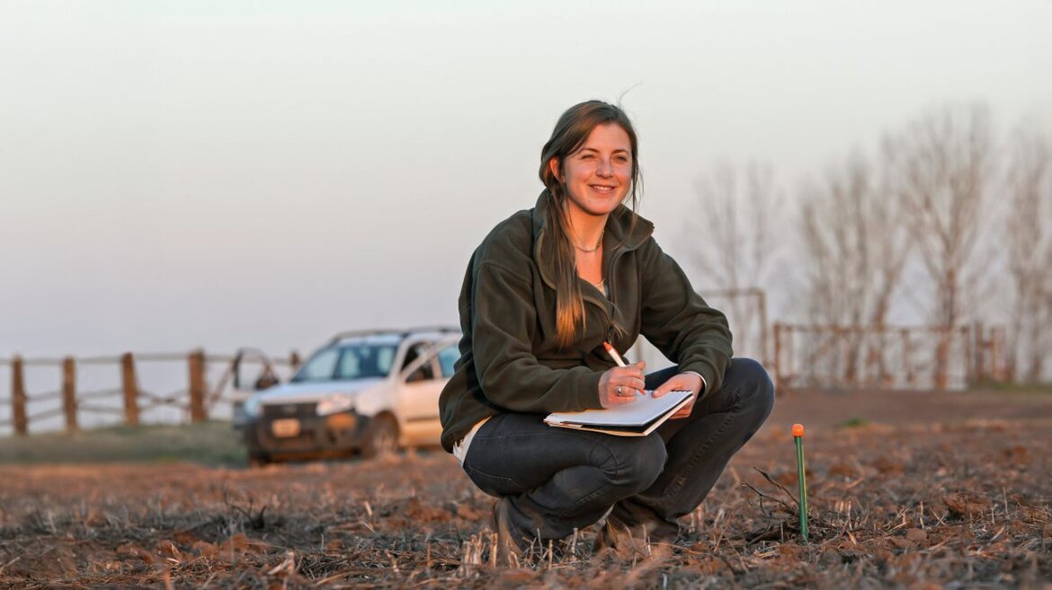 Frau mit Stift und Block hockt auf einem Feld und notiert Daten.