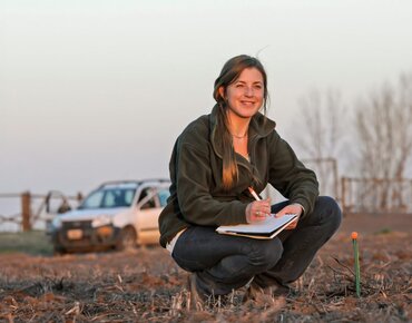 Frau mit Stift und Block hockt auf einem Feld und notiert Daten.