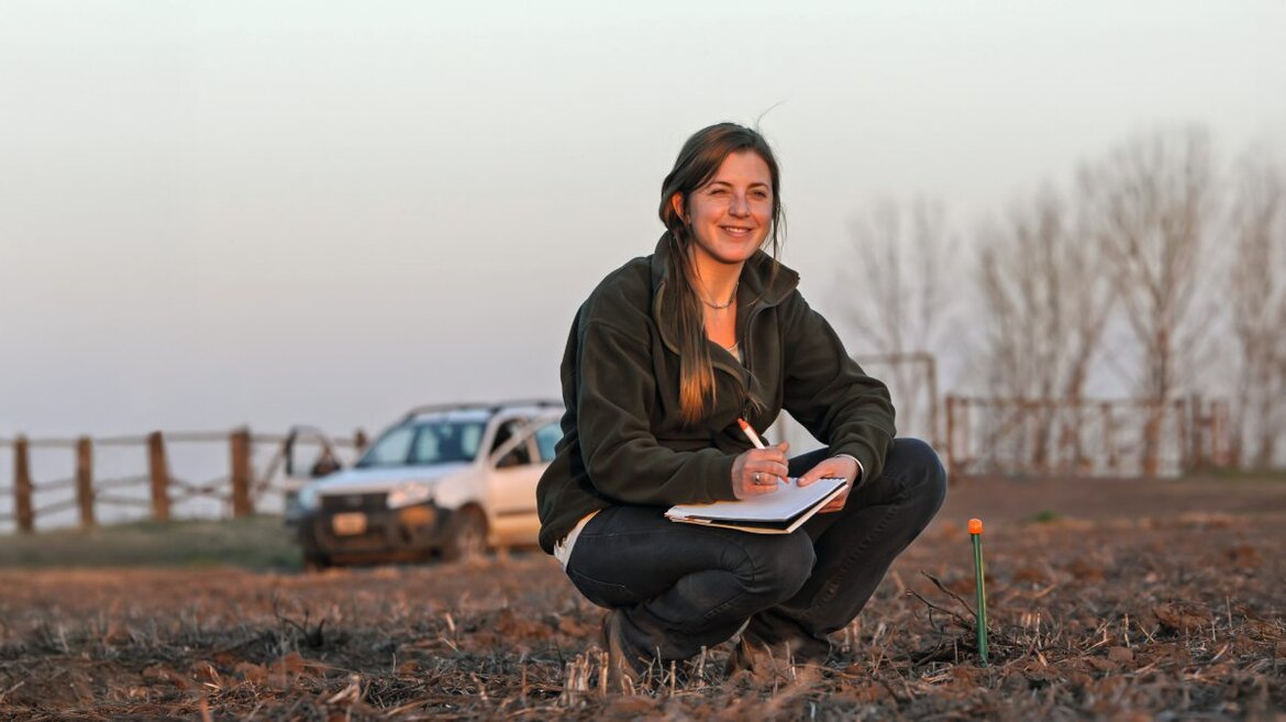 Eine junge Frau hockt mit einem Notizblock auf den Knien auf dem Feld